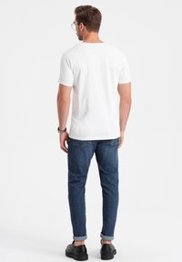 Hvid kortærmet t-shirt, blå jeans med oppressede cuff, sorte sko og et ur, set fra ryggen imod en ensfarvet baggrund.