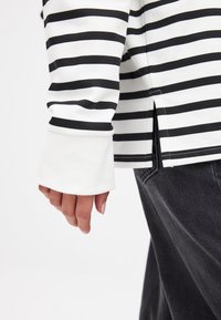 Črno-bela prugasta dolgo rokavna majica z belim manšetnim delom in stranskim razporkom. Tkanina se zdi mehka z gladko teksturo.