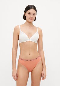 Fehér csipkéből készült háromszög alakú bralette vékony állítható pántokkal, korall színű hipster alsóneművel párosítva, fekete Calvin Klein logós derékpánttal.