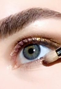 Primo piano di un occhio con un ombretto dorato scintillante applicato sulla palpebra, un morbido pennello in uso e sopracciglia definite con delicatezza sopra.