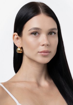 Goldene Chunky-Ohrringe mit einer glatten, polierten Oberfläche, die eine abstrahierte Form aufweisen, die nah am Ohr sitzt. Das Modell hat glattes schwarzes Haar.