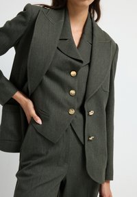 Donkergroene op maat gemaakte pak met een blazer met spitse revers, gouden knopen en een bijpassend vest. Soepele textuur met een gestructureerde pasvorm.