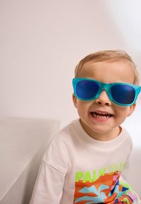 Helle türkisblaue Sonnenbrille mit blauen Gläsern, getragen von einem Kind. Das Sweatshirt ist hellfarben mit lebendigen grafischen Designs in Orange und Grün.