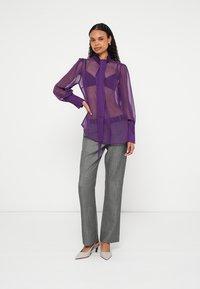 Διάφανη μοβ μπλούζα με μακριά μανίκια και δέσιμο στο λαιμό, συνδυασμένη με γκρι παντελόνι με σχέδια και ανοιχτό γκρι ψηλοτάκουνα παπούτσια.