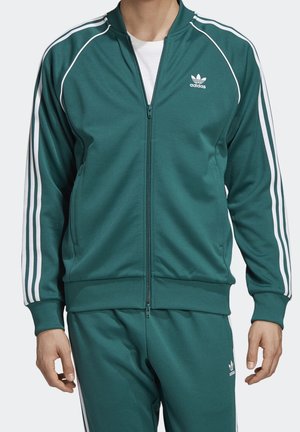 Homme portant un survêtement Adidas bleu sarcelle avec des bandes blanches sur les manches et les jambes, logos Adidas sur la poitrine et la cuisse, debout avec les bras détendus.