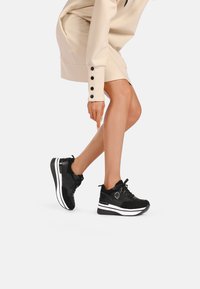Scarpe sportive nere con una combinazione di materiali lisci e testurizzati, una suola bianca a contrasto e lacci neri. Il design include un dettaglio con logo.
