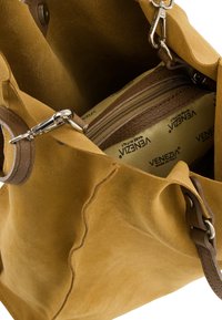 Sac en suède jaune moutarde avec des accents en cuir marron. L'intérieur révèle une pochette zippée avec un motif de logo sur un fond beige.