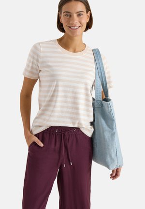 Uśmiechnięta kobieta w beżowo-paskowanej koszulce, bordowych spodniach z sznurkiem, niesie jasno-niebieską torbę na ramieniu na jednolitym tle.