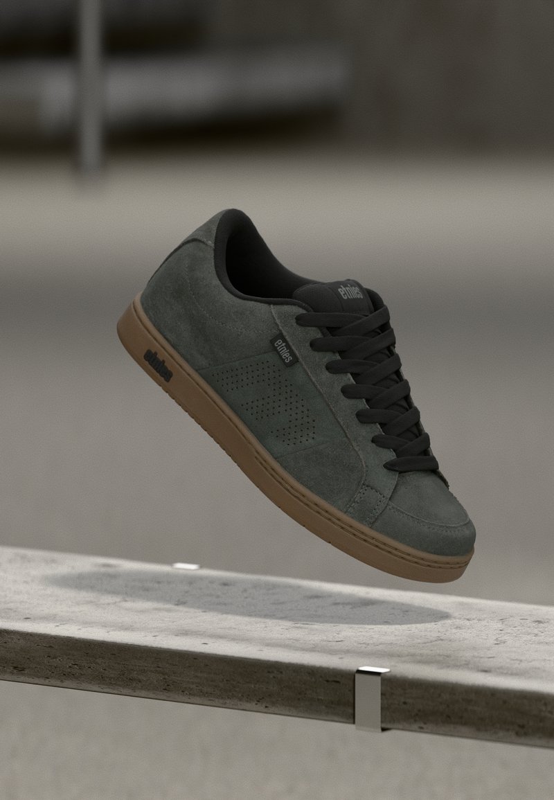 Green suede skate shoe with black laces, textile lining, perforated side panels, and a brown rubber sole. Floating above a surface.