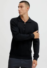 Black zip-up pullover in a textured fabric. Features a high collar and ribbed cuffs. Fitted design with a small branded accent at the hem.