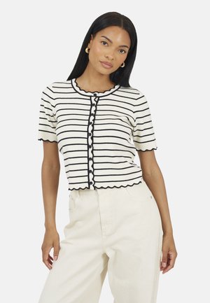 Kobieta o długich czarnych włosach, nosząca złote kolczyki koła, kremowo-czarną koszulę na krótki rękaw w paski zapinaną na guziki oraz kremowe spodnie z podwyższonym stanem.