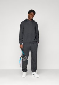 Mann i mørkegrå hettegenser og joggebukser som holder en tennisracket, står mot en enkel lys bakgrunn.