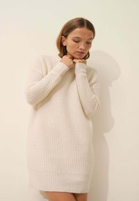 Dzianinowa sukienka w kolorze off-white, z teksturowanej tkaniny, długimi rękawami i prążkowanym wykończeniem. Prosty fason z luźnym krojem.