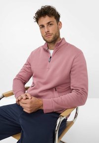Uomo con capelli ricci seduto su una sedia beige, indossa una felpa a mezza zip rosa e pantaloni blu scuro, con le mani incrociate in grembo, sfondo neutro.