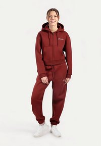 Sweat à capuche zippé couleur bordeaux avec poche kangourou et cordons de serrage, accompagné d'un pantalon de jogging assorti. Les deux présentent une texture douce et des poignets élastiques.