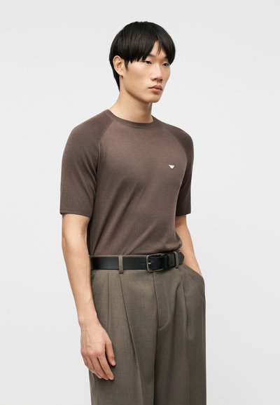 Νεαρός άνδρας με μαύρα μαλλιά που φοράει καφέ κοντομάνικο πουκάμισο και καφέ παντελόνι ψηλόμεσο με μαύρη ζώνη, στέκεται με τα χέρια στις τσέπες.