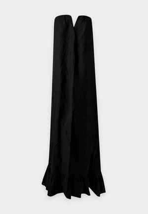 Une femme porte une robe noire bustier longue jusqu’au sol, à l’ourlet volanté, sur un fond clair et uni.