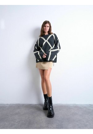 Jonge vrouw die een zwart-wit gebreide oversized trui draagt, een beige minirok en zwarte glanzende enkellaarsjes, staand tegen een witte muur.