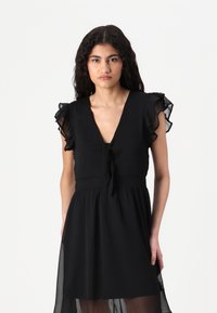 Black dress with a V-neckline, ruffled short sleeves, and a tie at the waist. Sheer fabric at the hem adds contrast to the solid design.
