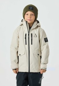 Beige waterproof jacket with a hood, featuring front zip and pockets, black accents, and logo details. Worn with a green knit beanie.