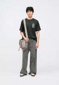 Tricou negru cu mâneci scurte și un imprimeu colorat, blugi gri lejeri și o geantă de umăr gri cu mai multe buzunare. Adidași maro și albi.