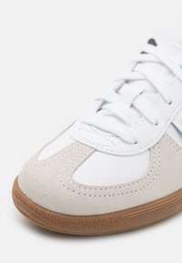 Balsvos sportiniai bateliai su suede smėlio spalvos akcentais, tekstūruota guma padais ir baltais raišteliais. Savybės - siuvimo detalės palei siūles ir minimalus dizainas.