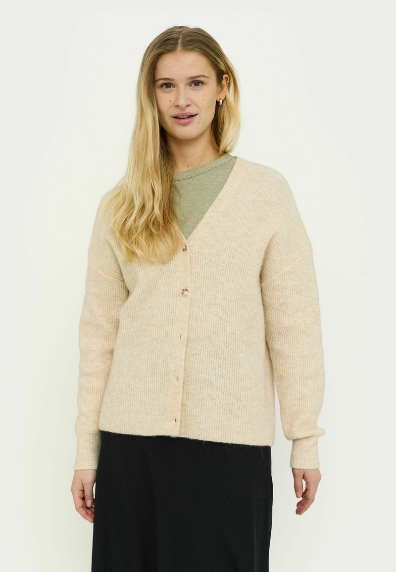 Cardigan beige clair avec un col en V, texture côtelée et cinq boutons sur le devant. Porté sur une chemise verte et associé à une jupe noire.