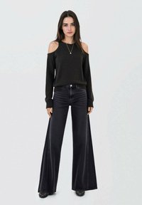 Černý pletený svetr s odhalenými rameny, v kombinaci s džínami se zvýšeným pasem a širokými nohavicemi v tmavém denimu. Modelka má na sobě černé boty a jednoduchý náhrdelník.