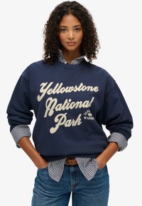 Sudadera azul marino con el texto "Parque Nacional Yellowstone" en color crema, con mangas largas y un cuello de canalé, superpuesta sobre una camiseta de rayas.