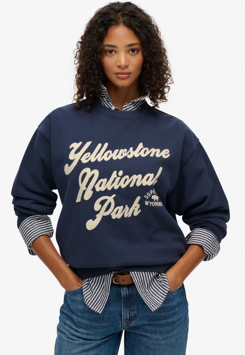 Sudadera azul marino con el texto "Parque Nacional Yellowstone" en color crema, con mangas largas y un cuello de canalé, superpuesta sobre una camiseta de rayas.
