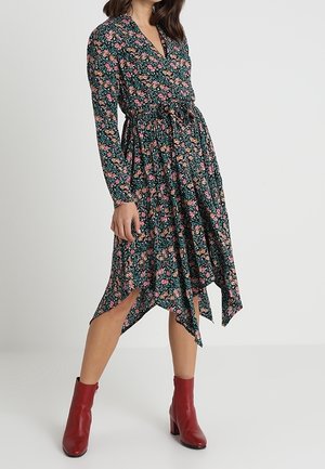 Robe portefeuille florale à manches longues, fond vert, fleurs roses et jaunes, ourlet asymétrique, taille cintrée, associée à des bottines rouges.