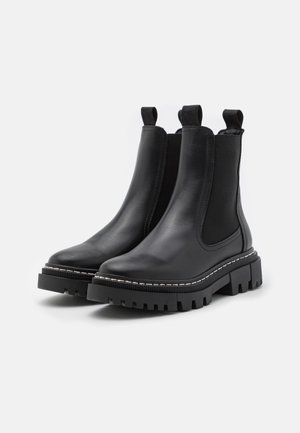 Botas Chelsea de cuero negro con lados elásticos, tiradores y suelas de goma con tacos gruesos. Presenta costuras blancas a lo largo del borde de la suela.