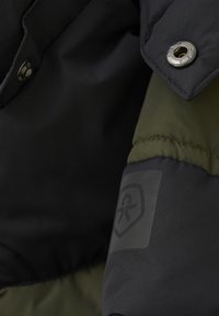 Detalj av en svart och olivgrön isolerad jacka med tryckknapp, slät yta och en logotyp på ärmen.