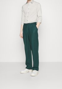 Camisa de botones de color gris claro, pantalones verdes con pliegues y zapatos blancos con suela gruesa. La camisa tiene las mangas remangadas y un corte relajado.