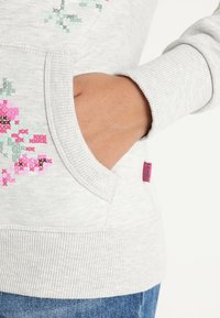 Sudadera gris con bordado floral en rosa y verde. Presenta un bolsillo lateral y puños acanalados. Etiqueta de la marca en rosa visible.