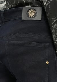 Fekete farmernadrág hátsó zsebében arany gomb található, felette pedig Versace Jeans Couture fekete bőr címke látható, amit fekete mintás inggel viselnek.
