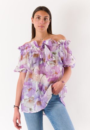 Donna che indossa una blusa scollata con stampa floreale viola e jeans azzurri, in piedi con una mano in tasca su sfondo bianco.