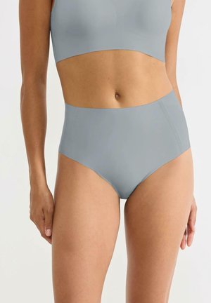 Kobieta w jasnoniebieskich, wysokich majtkach i dopasowanym sportowym staniku, stoi na jednolitym białym tle.