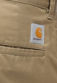 Tkanină de culoare nisipie, cu o textură fină, având o pată albă cu un logo portocaliu și detalii cusute, situată pe un buzunar al unui articol de îmbrăcăminte.