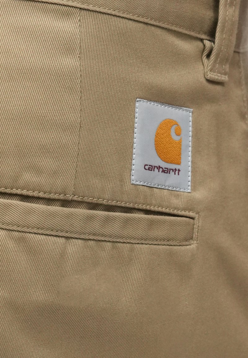 Beige tyg med en slät struktur, som har en vit lapp med en orange logotyp och sydda detaljer, placerad på en ficka av plagget.