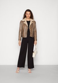 Brun skinnjakke med shearling-krage, svart topp, svarte vide bukser, hvit håndveske og hvite sandaler med remmer mot en ensfarget bakgrunn.
