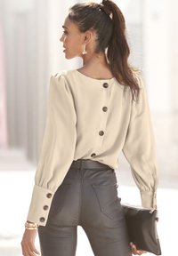 Beige Bluse mit Puffärmeln und Knopfakzenten auf der Rückseite, kombiniert mit hoch taillierten schwarzen Hosen und einer schwarzen Clutch in der Hand.