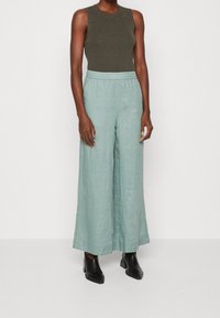 Jasnozielone lniane spodnie o szerokiej nogawce z elastycznym paskiem, zestawione z dopasowanym, ciemnozielonym topem bez rękawów i czarnymi botkami na obcasie.