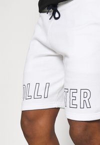 Vita bomullsshorts med lös passform, dragsko i midjan och stora tryckta bokstäver på vänster ben. Slät textur med minimal design.