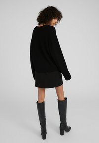Schwarzer, gerippter Pullover mit lockerer Passform, kombiniert mit einem kurzen schwarzen Rock. Das Model trägt hohe schwarze Lederstiefel mit Blockabsätzen.