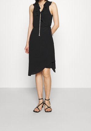 Zwarte mouwloze jurk met geruchte halslijn en asymmetrische zoom, gemaakt van lichtgewicht stof. Gecombineerd met zwarte sandalen met bandjes.