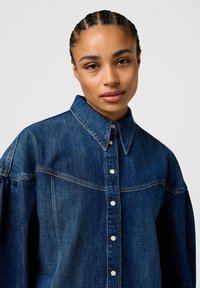 Chemise en denim bleu foncé avec un col pointu, fermeture à boutons à l'avant avec des boutons en métal, et des manches bouffantes. Texture lisse et design décontracté.