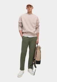 Hellbeige Sweatshirt, grüne Hose, weiße Sneakers, beiger Beutel mit schwarzen Akzenten. Das Modell posiert vor einem weißen Hintergrund.