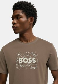 Camiseta de manga corta marrón con un estampado texturizado en blanco que presenta la palabra "BOSS" rodeada por un patrón mármol.
