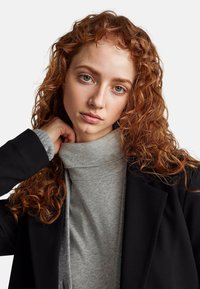 Černý blazer s měkkým šedým rolákem. Texturované kudrnaté červené vlasy přidávají hloubku. Neutrální pozadí zvýrazňuje detaily oblečení.